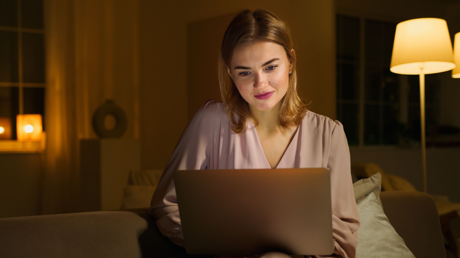 Une jeune femme souriante travaille sur son ordinateur portable dans un salon chaleureux, éclairé le soir par une lampe.